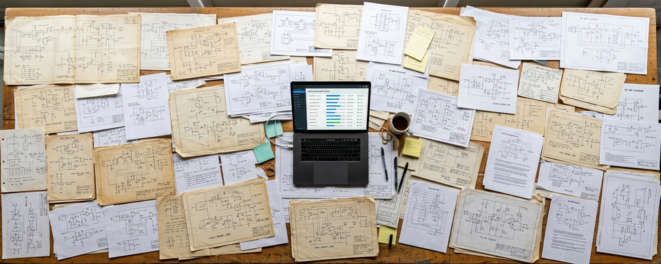 Dozens of schematics spread on table with laptop processing queue